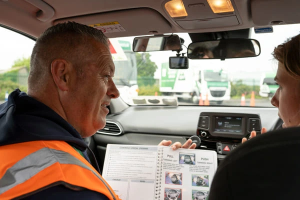 A_driving_instructor_in_an_orange_vest_speaks_to_a_woman_seated_in_the_car_ISM_Driving_lessons