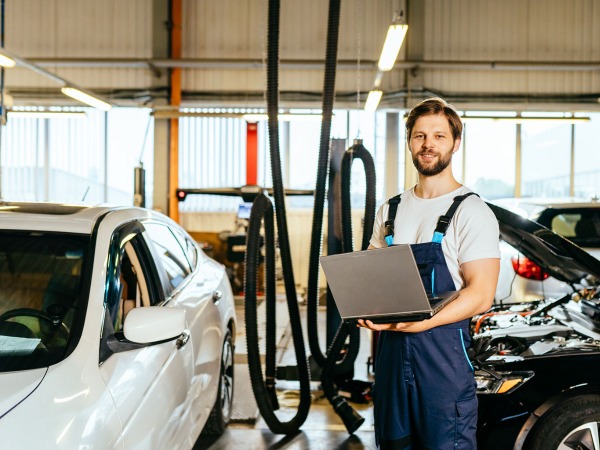 A_man_in_overalls_holds_a_laptop_in_front_of_a_parked_car_ISM_Driving_lessons